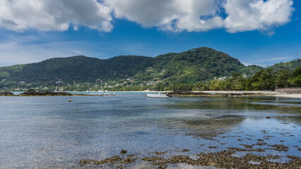 Low tide on a tropical beach. The exposed bottom is visible through the clear ocean water. The boat is moored in shallow water. Coastal green hill on the background of blue sky and clouds. Seychelles