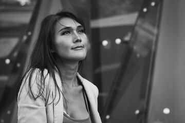Hopeful look of Thai businesswoman outside glass office building in urban city. Pensive transgender Asian person thinking about project ideas. Black and white photography
