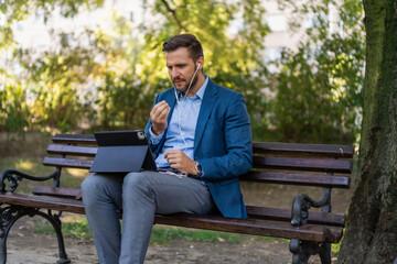 Obraz premium Caucasian male entrepreneur freelancer sitting outdoors in city park a bench talking online a video call using laptop or tablet. Business man office worker working on urban street background 