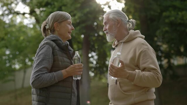 Mature lovely family drinking water, talking each other prepare for doing sport standing in the part. Elderly couple feeling thirsty. Couple of pensioners. People and communication concept.