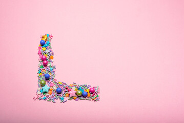 Sparkles, sprinkles and small multicolored sugar spheres forming a letter L on a pink background.