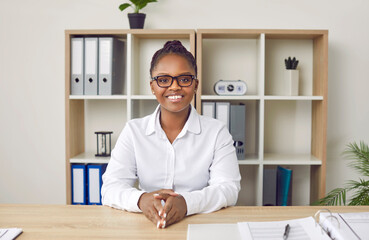 Happy beautiful young African American woman in white shirt and eyeglasses sitting at office desk,...