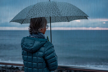 Back view of senior woman at sea in a rainy evening holding an umbrella while strolling outside....