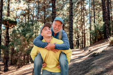 Handsome boy carries his grandfather on shoulders as he walks happily in the forest. Smiling multigeneration family couple enjoying mountain and nature together. The new generation helps the old one