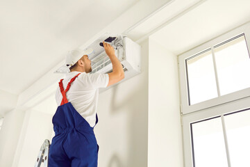 Male technician repairs, cleans or installs air conditioner hanging on wall inside house. Man in...