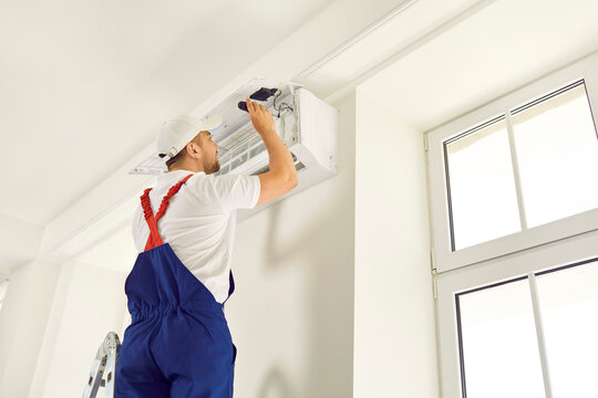 Male technician repairs, cleans or installs air conditioner hanging on wall inside house. Man in work clothes and works on call at client's house. Concept of service and maintenance of air conditioner