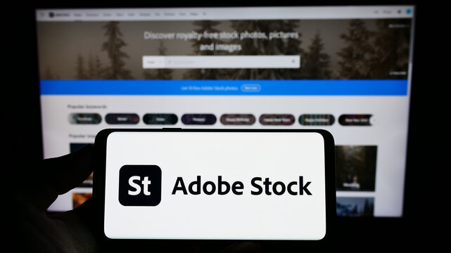 Stuttgart, Germany - 12-30-2022: Person holding mobile phone with logo of American microstock agency Adobe Stock on screen in front of business web page. Focus on phone display.