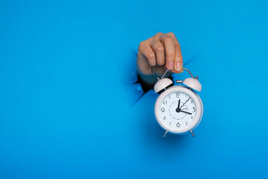 female hand holds pink alarm clock through a paper hole in blue background.
