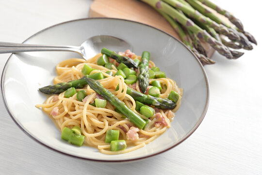 Spaghetti with green asparagus, bacon and egg as carbonara sauce variation on a plate and on a white painted wooden table, selected focus