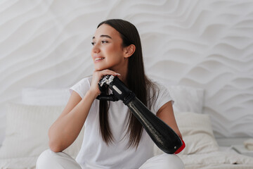 Close up of pretty young woman lean on her bionic cyber hand, looks aside grateful for full life. Beautiful American girl with artificial arm. Cybernetic technologies, medicine for people after trauma