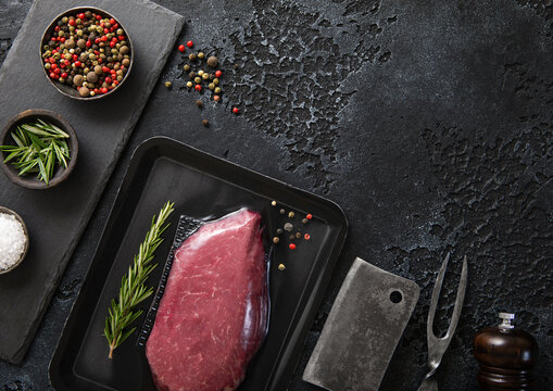 Raw beef fillet steak in vacuum tray with pepper on on black background with meat cleaver and fork.