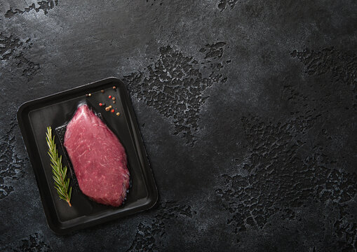 Fresh raw beef fillet steak sealed in vacuum tray with pepper and rosemary on black kitchen table background.