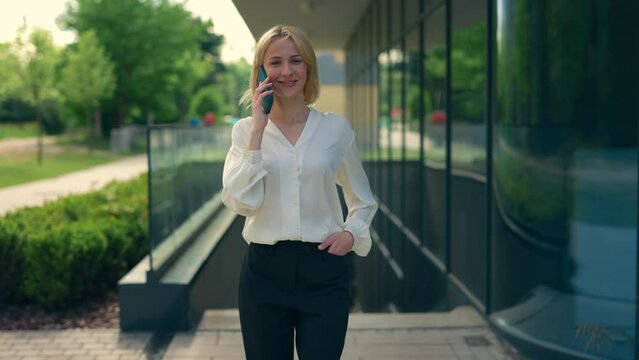 Happy business woman in formal suit going near the business centre speaking on the phone with clients. Female manager talking on cellphone outside. People and mobile communication.
