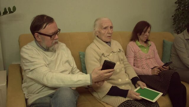 Family members use smartphones, tablet pc ( green screen ) and laptop.  Slow motion 2x.  An elderly man, a grandmother, a middle-aged woman and a young woman are sitting on a sofa in a room. 