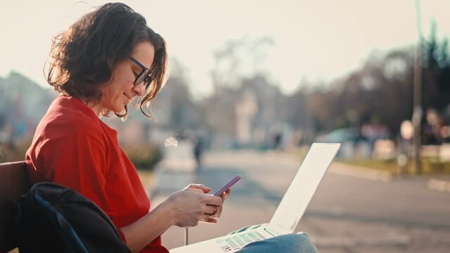 Young caucasian woman in glasses working or studying using a laptop and smartphone while sitting on a bench outdoors
