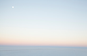 An empty scene of white salt flat and pale, early sunrise sky with the moon in top left corner.