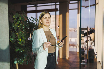 A successful young woman in glasses and a jacket is standing in the office and happily talking on the phone. A young manager works in the office and manages the business remotely