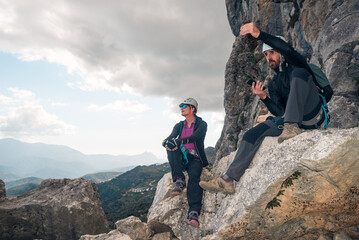 Concept: adventure. Man and woman climbers with helmet and harness. Resting sitting on a rock. Talking and gesturing while looking at the horizon. Via ferrata in the mountains.