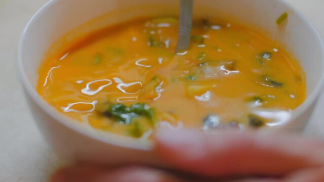 Woman stirs tasty and spicy Asian Tom Yam soup. Bright orange hot soup with meat and vegetables in porcelain bowl.