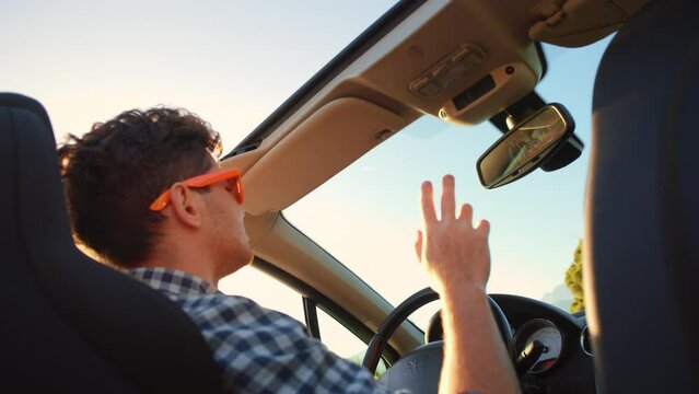 Young stylish man in sunglasses driving cabriolet car waving and dancing hand at golden sunset. Concept of success, tourism and rental. Happy smiling male tourist travel in California. Summer vacation