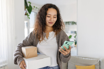 Smiling young Asian woman shopping online with smartphone on hand, receiving a delivered parcel by...