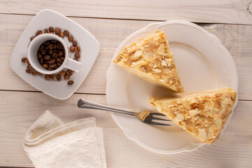 Two pieces of cream cake with a white saucer, a metal fork and a cup with coffee beans on a wooden table, macro, top view.