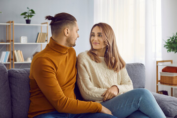 Portrait of a young couple looking to each other at home. Happy romantic spouses wearing knitted pullovers hugging while sitting on couch in living room. Love, relationship concept