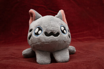 A toy plush cat in gray lies on a dark red background.