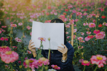 Female covering her face with blank book or magazine with zinnia flowers in a garden