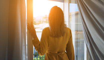 Woman enjoys warm summer breeze while standing in front of the window.