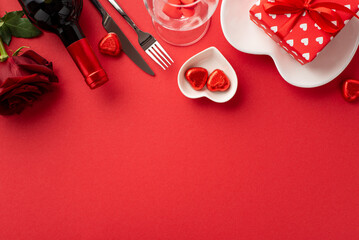 Valentine's Day concept. Top view photo of wine bottle glass giftbox rose heart shaped plates cutlery and chocolate candies on isolated red background with copyspace