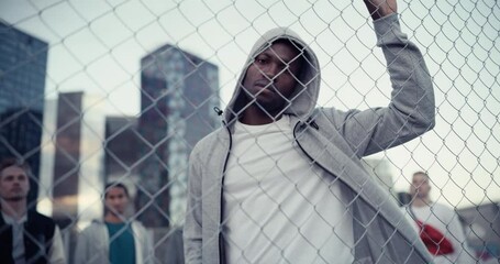 Portrait of a Handsome Athletic Black Man in a Hoodie Posing and Looking at Camera while Standing Behind a Fence on a Rooftop Urban Parking Lot. Diverse Friends Standing Behind Him - Powered by Adobe