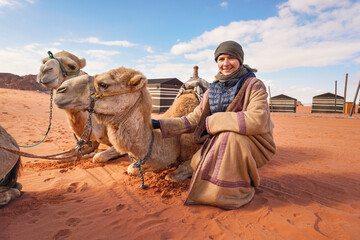 Fototapeta premium Young woman in traditional Bedouin coat - bisht - and headscarf crouching next to two camels laying on red desert ground, smiling, camp tents in background