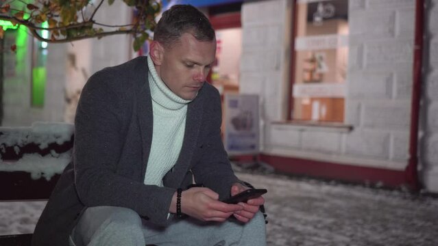 Handsome man scrolling social media on mobile phone sitting outdoors at cold snowy winter at night. Posting social media, online shopping, texting, e commerce