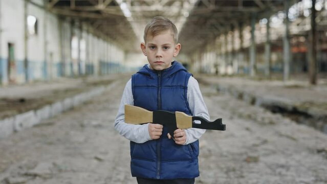 Child holds a toy gun standing against a background of devastation looking at camera. Portrait. Boy games. War. Boy playing in war