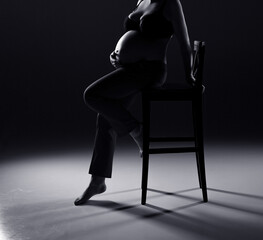 A pregnant woman is sitting on a chair on a gray background. Silhouette of a pregnant woman. Black and white photo. Studio pregnancy photo shoot. Woman sitting on chair.