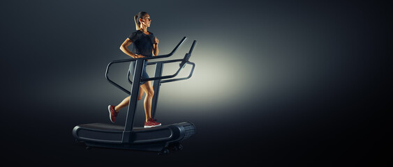 Full length profile shot of a young sporty female athlete running on a treadmill isolated on dark background, Conceptual wide banner studio photo. Training at home.