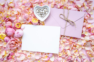 Background of pink rose buds and petals with envelope and blank card in centre and ceramic plate with wooden heart. Flat lay, overhead view. Mother's Day, St. Valentine's Day, March 8 concept.