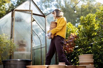 Woman stands with a pile of clay jugs in front of glass orangery at garden in sunny morning. Gardening and hobby concept - obrazy, fototapety, plakaty