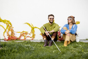 Young and stylish couple of friends sit together on white wall background with ivy outdoors, hanging out together and having fun
