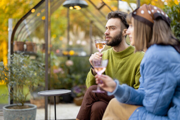 Young stylish couple hang out together, talk and drink wine while sitting at backyard. Man and woman spending evening time at cozy atmosphere outdoors