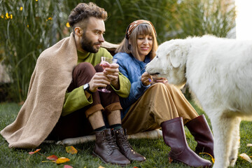 Young stylish couple warming up covered with plaid while sitting together and feed their cute dog on green lawn at backyard. Young family spending autumn time outdoors