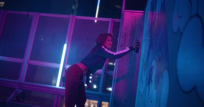 Young Beautiful Woman is Creating a Graffiti Masterpiece on a Wall at a Parking Lot with Neon Lights at Night. Athletic Female Using Spray Can to Release Her Creative Energy