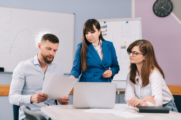 A team of business people are discussing the project together in the office. The team sits at a table and looks at a laptop screen.