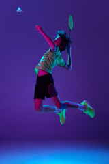 Fototapeta na wymiar Concentrated teen boy in uniform playing badminton, hitting shuttlecock in a jump over blue purple background in neon ligth