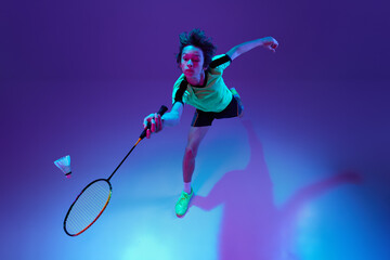 Top view. Portrait of teen boy in uniform playing badminton, returning shuttlecock in a run over blue purple background in neon ligth
