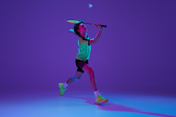 Fototapeta na wymiar Dynamics. Portrait of teen boy in uniform playing badminton, hitting shuttlecock in a run over blue purple background in neon ligth