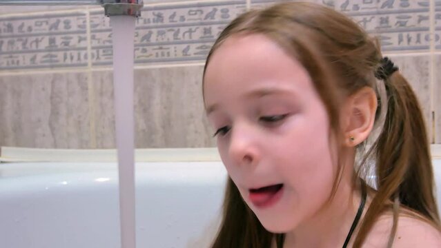 A little girl sitting in the bathroom is washing her face nicely, taking water in her hands, rubbing her face, pouting her lips, hair is wet and disheveled. The concept of children's oral hygiene.