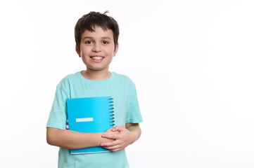 Adorable smart teen schoolboy in blue shirt holding a copybook and smiling a cheerful toothy smile looking at camera, isolated on white background with copy space. Back to school concept. Education.