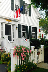 A house in the summer with a garden and flowers blooming hangs a flag in front for the Fourth of...